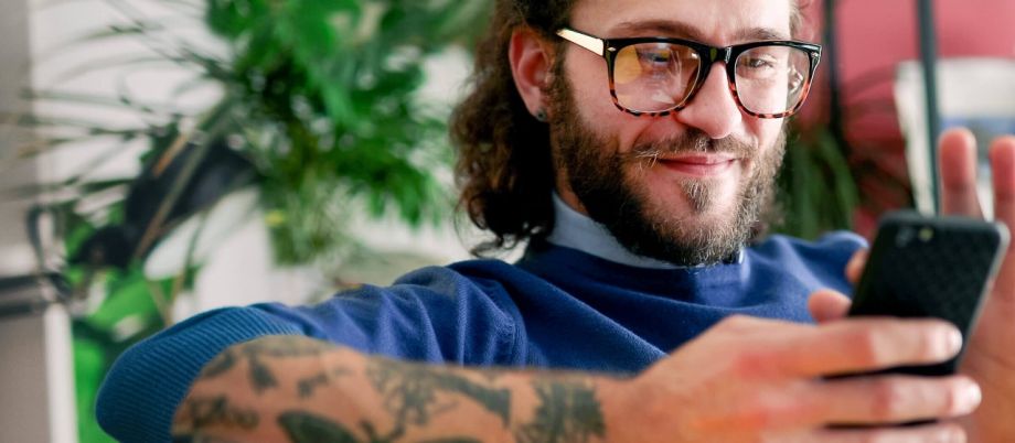 Mann mit Brille, Tattoos und Bart schaut auf sein Handy